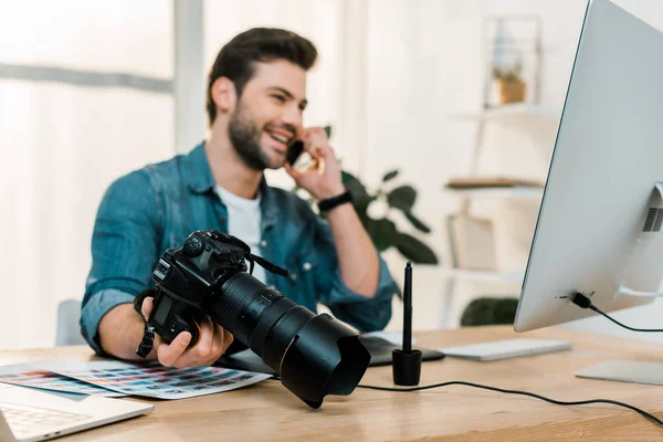 Jovem fotógrafo feliz segurando câmera e falando por smartphone na mesa de trabalho — Fotografia de Stock