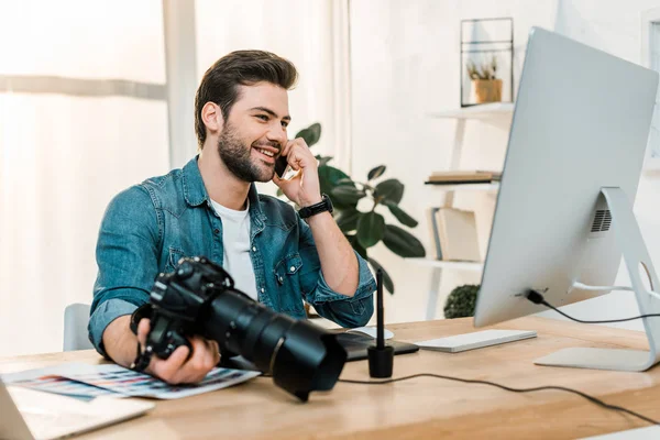 Улыбающийся молодой фотограф держит камеру и разговаривает со смартфоном в офисе — стоковое фото