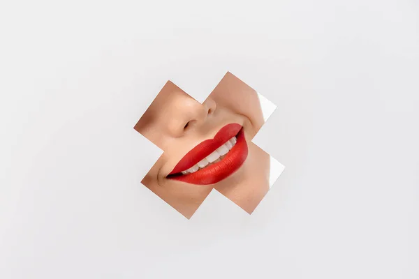 Cropped shot of smiling young woman with red lips through cross shaped hole on white — Stock Photo