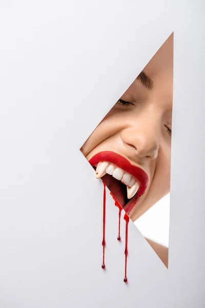 Cropped shot of woman with vampire teeth and blood biting through hole on white — Stock Photo