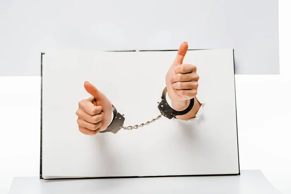 Partial view of female hands in handcuffs showing thumbs up through holes on white — Stock Photo