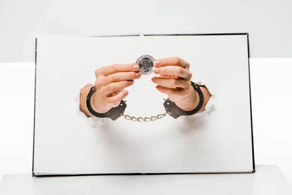 Cropped shot of woman in handcuffs holding bitcoin through holes on white — Stock Photo