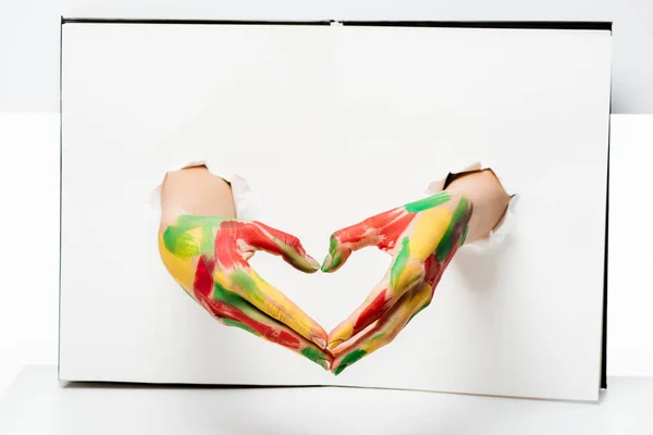 Cropped shot of woman with hands in paint showing hand heart symbol through holes on white — Stock Photo