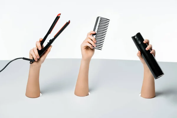 Cropped shot of women holding hair spray, comb and hair straightener through holes on white — Stock Photo