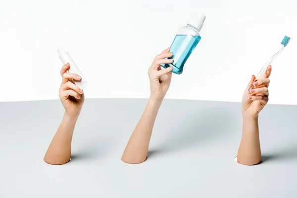 Visão parcial de mulheres segurando pasta de dentes, escova de dentes e enxaguatório bucal através de furos no branco — Fotografia de Stock