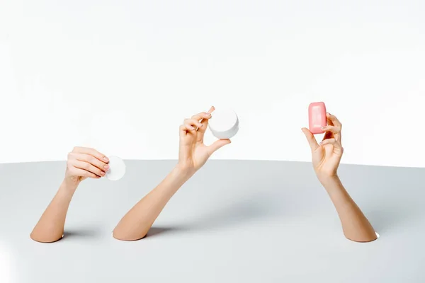 Tiro recortado de manos femeninas sosteniendo jabón, almohadilla de algodón y crema a través de agujeros en blanco - foto de stock
