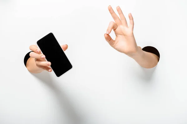 Cropped image of woman holding smartphone with blank screen and showing okay gesture through holes on white — Stock Photo