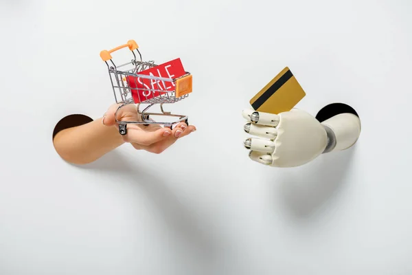 Imagen recortada de mujer y robot sosteniendo pequeño carro de la compra y tarjeta de crédito a través de agujeros en blanco - foto de stock