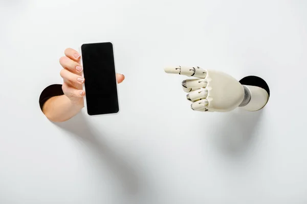 Imagen recortada de la mujer sosteniendo teléfono inteligente con pantalla en blanco y robot apuntando a través de agujeros en blanco — Stock Photo
