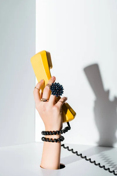 Imagen recortada de la mujer sosteniendo el auricular amarillo de teléfono retro en la mano a través del agujero en blanco - foto de stock