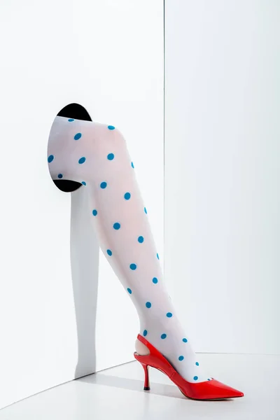 Cropped image of girl showing leg in fashionable white tights with blue dots and red high heel in hole on white — Stock Photo