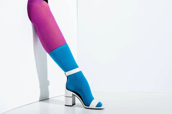 Обрізане зображення дівчини, що показує ногу в модних фіолетових колготках, синій шкарпетка і білий високий каблук в дірі на білому — стокове фото