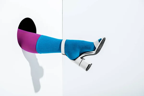 Cropped image of girl showing leg in trendy violet tights, blue sock and white high heel in hole on white — Stock Photo