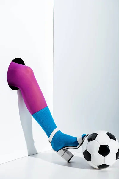 Abgeschnittenes Bild eines Mädchens, das ein Bein in violetten Strumpfhosen, blauer Socke und weißem High Heel auf einem Fußballball durch ein Loch auf weißem Grund steckt — Stockfoto