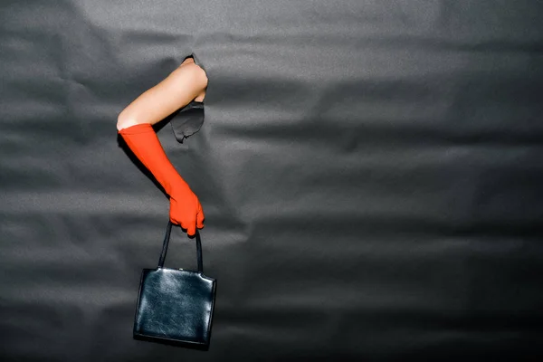 Immagine ritagliata di ragazza che mostra mano in guanto arancione e borsetta nera attraverso carta nera — Foto stock