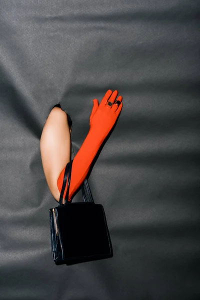 Imagem cortada de menina mostrando mão na luva laranja elegante e bolsa preta através de papel preto — Fotografia de Stock