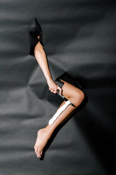 Imagem cortada de menina perna de barbear com faca através de furos em papel preto — Fotografia de Stock
