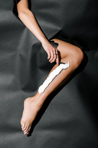 Imagen recortada de la pierna de afeitar chica con cuchilla a través de agujeros en papel negro - foto de stock