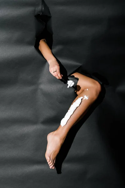 Imagen recortada de la pierna de afeitar chica con cuchilla a través de agujeros en papel negro - foto de stock