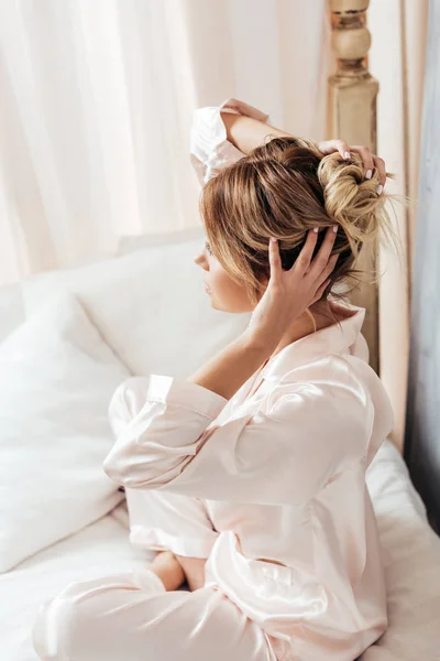 Vista lateral de la mujer en pijama ajustando el pelo en la cama durante la mañana en casa - foto de stock