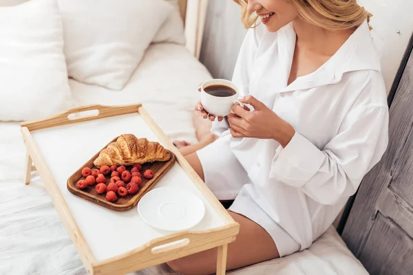 Частковий вид усміхненої дівчини, що тримає чашку кави, сидячи на ліжку зі сніданком у підносі — стокове фото