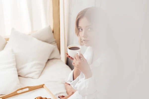 Foco seletivo da jovem segurando xícara de café e tomando café da manhã na cama — Fotografia de Stock