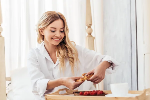 Felice donna strappando croissant a letto con la prima colazione sul vassoio durante il mattino a casa — Foto stock