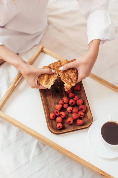 Imagen recortada de la mujer rompiendo croissant en la cama con desayuno en bandeja durante la mañana en casa - foto de stock