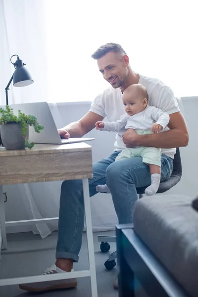 Glücklicher Vater sitzt auf Stuhl, hält Baby und benutzt Laptop zu Hause — Stockfoto