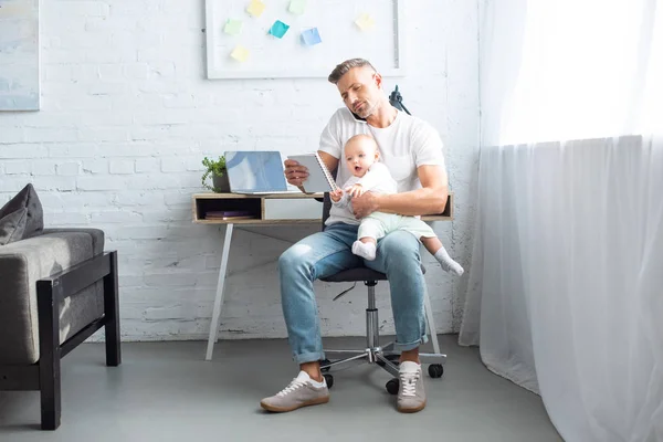 Father sitting on chair, looking at notebook, holding adorable baby daughter and talking on smartphone at home — Stock Photo