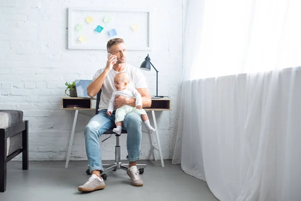 Vater sitzt auf Stuhl, telefoniert mit Smartphone und hält kleine Tochter im Wohnzimmer — Stockfoto