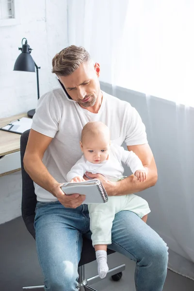 Schöner Vater sitzt auf Stuhl, telefoniert mit Smartphone, schaut auf Notizbuch und hält entzückende kleine Tochter zu Hause — Stockfoto