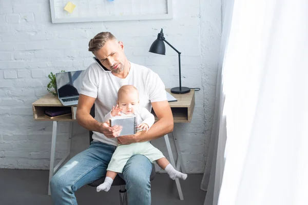 Busy father sitting on chair with notebook, talking on smartphone and holding baby daughter at home — Stock Photo