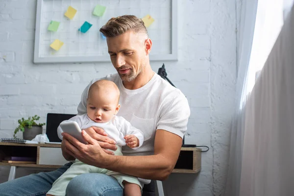 Padre sentado con adorable hija bebé en la silla y el uso de teléfono inteligente en casa - foto de stock