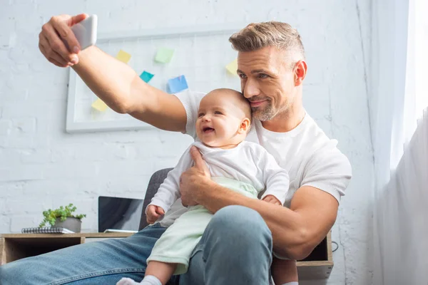 Padre felice seduto sulla sedia, tenendo in braccio la bambina e scattando selfie sullo smartphone a casa — Foto stock