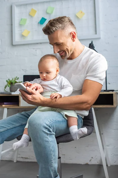 Pai sentado na cadeira, segurando adorável bebê filha e usando smartphone em casa — Fotografia de Stock