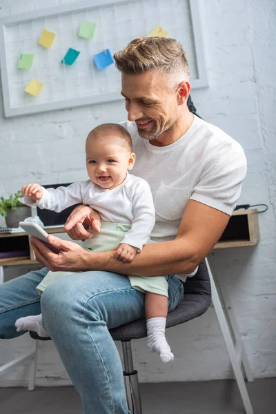 Glücklicher Vater sitzt auf Stuhl, hält entzückende kleine Tochter und zeigt zu Hause mit dem Finger auf Smartphone — Stockfoto