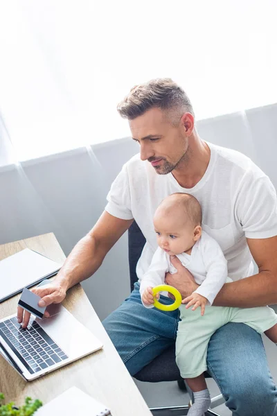 Padre guapo sentado en el escritorio de la computadora, haciendo compras en línea con tarjeta de crédito y sosteniendo a la hija del bebé en casa - foto de stock