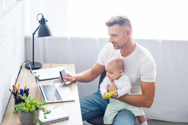 Handsome father sitting at computer desk with credit card for online shopping and holding baby daughter at home — Stock Photo