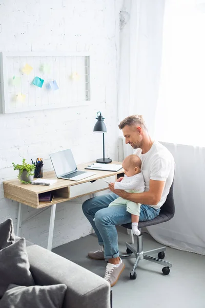 Padre sentado en el escritorio de la computadora, usando un teléfono inteligente y sosteniendo a la hija en la sala de estar - foto de stock