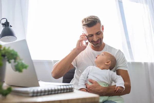 Happy father sitting at computer desk, talking on smartphone and holding baby daughter at home — Stock Photo
