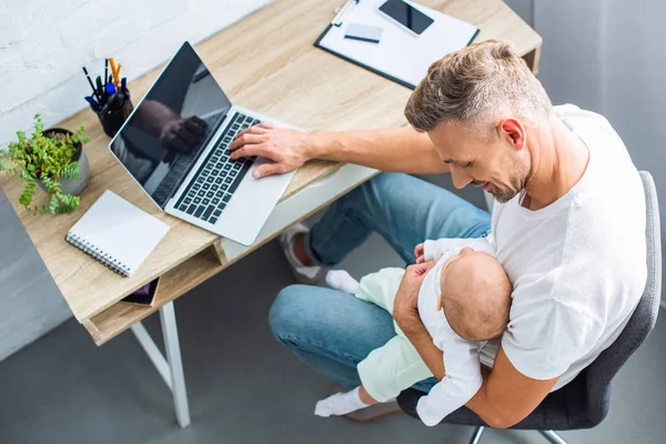 Father sitting at desk and using laptop with blank screen while holding baby daughter at home — Stock Photo