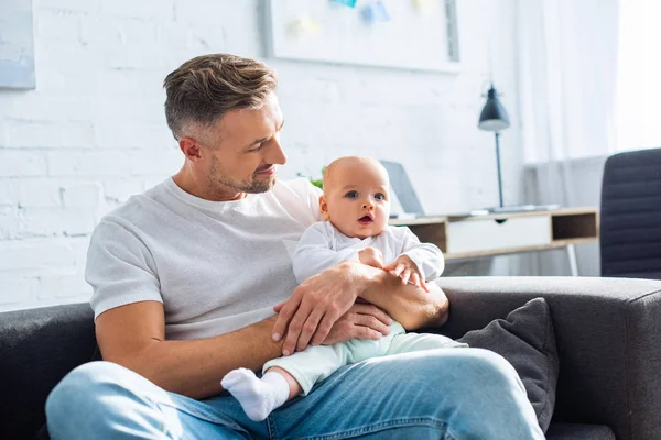 Handsome smiling father sitting on sofa and holding adorable baby daughter at home — Stock Photo