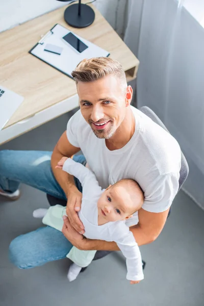 Smiling father sitting on chair, holding baby daughter and looking at camera — Stock Photo