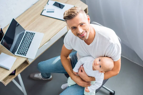 Padre sentado en el escritorio de la computadora, sosteniendo a la hija del bebé y mirando a la cámara en casa - foto de stock