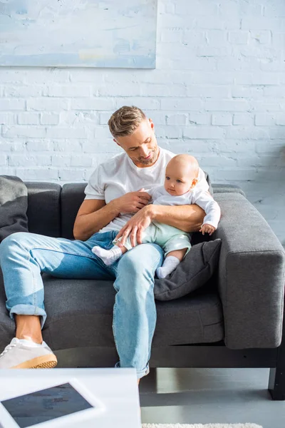 Bonito pai sentado no sofá e segurando bebê filha na sala de estar — Fotografia de Stock