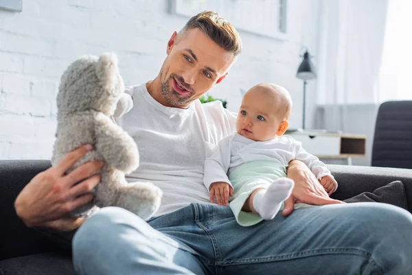 Padre sentado en el sofá con adorable hija bebé y jugando con el oso de peluche en casa - foto de stock