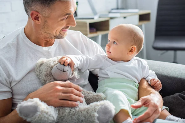 Adorable bébé fille jouer avec ours en peluche tout en étant assis avec le père souriant sur le canapé à la maison — Photo de stock