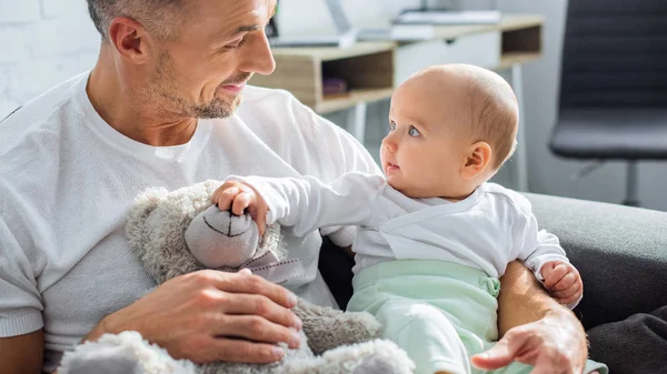 Adorable bebé hija jugando con osito de peluche mientras sentado con padre en sofá en casa - foto de stock