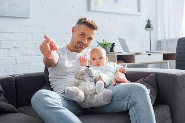 Smiling father sitting on couch with baby daughter and pointing with finger at something — Stock Photo